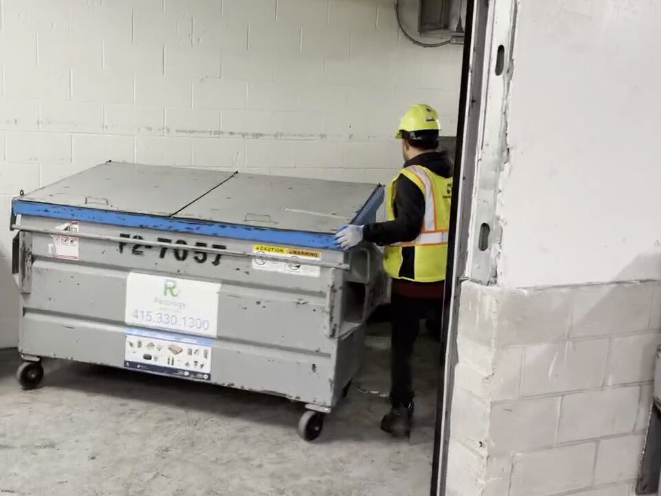 An Ally Waste team member in a hard hat and reflective vest rolls a dumpster across a building's trash room