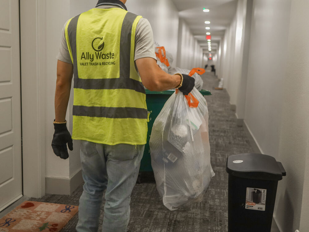 An Ally Waste valet in a reflective vest carries a resident's trash bag down an apartment hallway to a collection cart