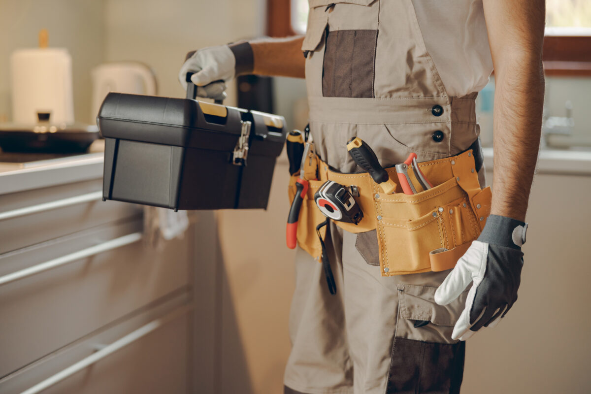 Close up of repairman in uniform standing on home kitchen and holding his tool bag | Ally Waste