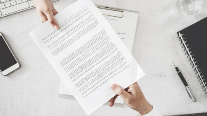 Two hands hold a printed page over a light desk with a notebook, pen, glass, phone, and keyboard nearby.