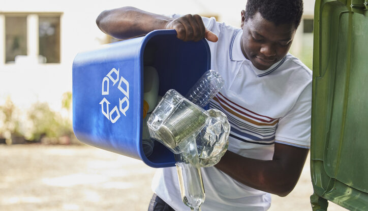 A man dumps plastic bottles from a blue recycling bin into a large green outdoor recycling container.