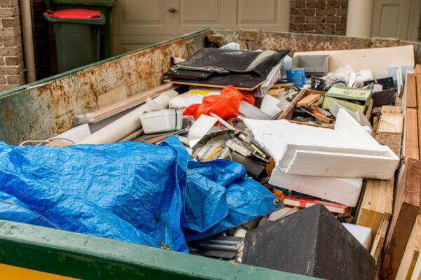 Open dumpster overflowing with construction debris: wood, foam, metal pieces, and a blue tarp covering part of the pile in front of a home.