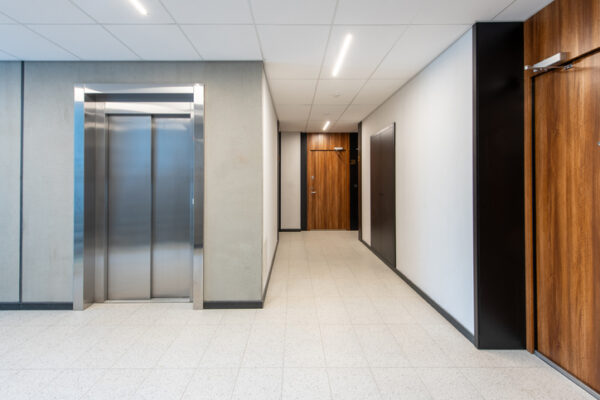 Modern office hallway featuring an elevator to the left and wooden doors along a bright corridor.