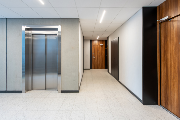 Modern office hallway featuring an elevator to the left and wooden doors along a bright corridor.