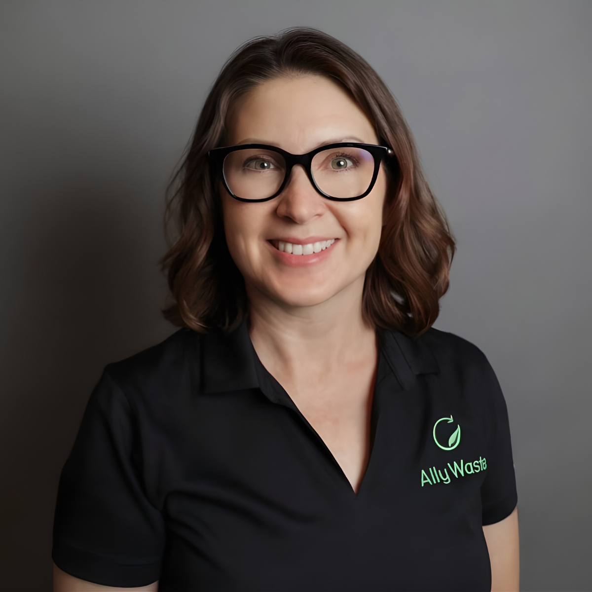 Portrait of a smiling woman with short brown hair and black glasses, wearing a black polo with a green Ally Waste logo against a gray backdrop.
