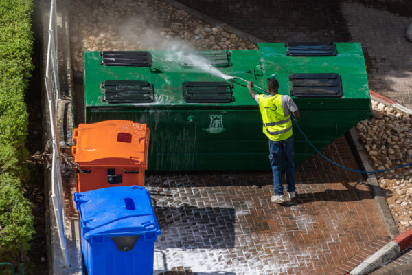 Sanitation worker in a yellow vest pressure-washes a large green dumpster outside, with orange and blue waste bins nearby.