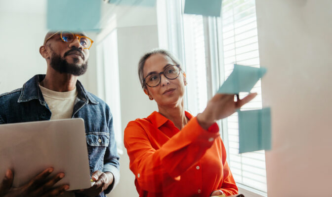 Two colleagues brainstorming in an office, woman in orange blouse placing sticky notes on a glass board while a man with a tablet watches.