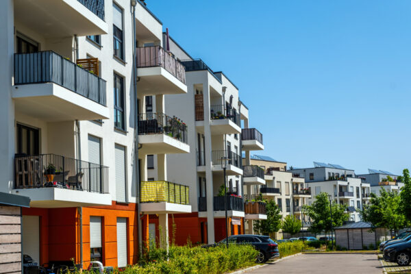 Row of modern white apartment buildings with open balconies and orange ground floors under a clear blue sky; a street with parked cars and greenery runs alongside the buildings.